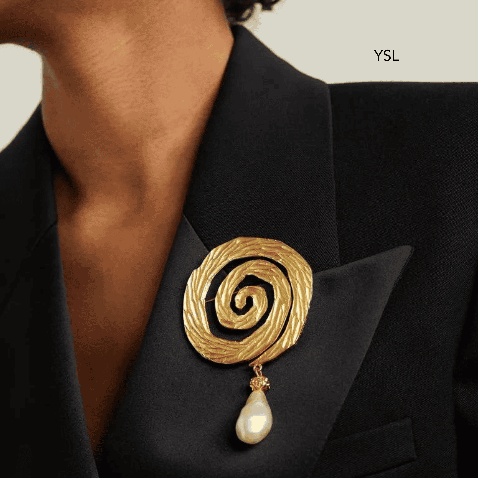 Close-up of a black tailored blazer featuring a statement gold spiral brooch with a textured finish and a dangling teardrop pearl. The brooch is pinned to the blazer's lapel, adding an elegant, vintage-inspired accent to a sophisticated outfit.