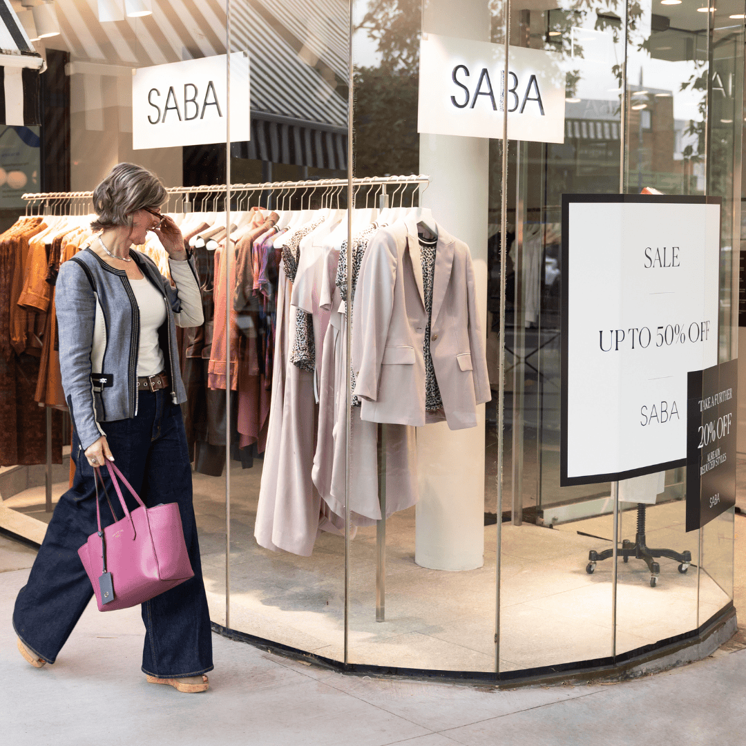 A woman carrying a pink handbag looks toward a clothing store window featuring a sale sign advertising up to 50% off.